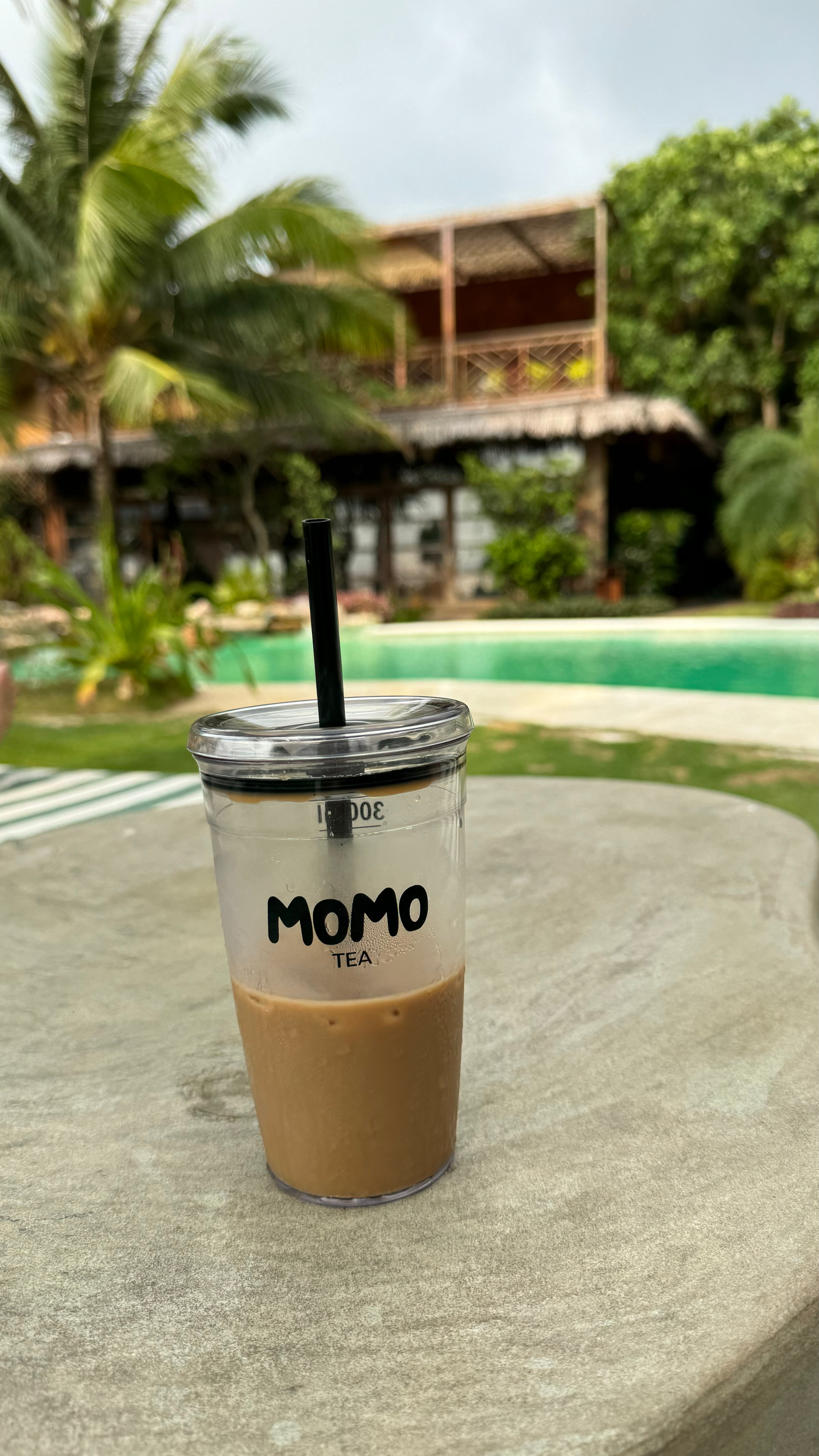 Clear cup with iced coffee and straw on a stone surface, pool and tropical trees in the background