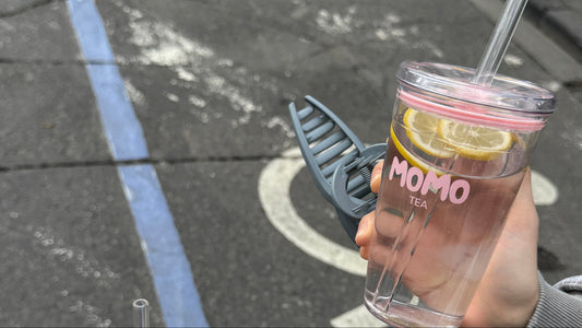 Two hands holding clear plastic cups with straws on an outdoor street. The right cup has a pink lid and lid rim, a lemon slice, and the word 'MOMO' printed on it.
