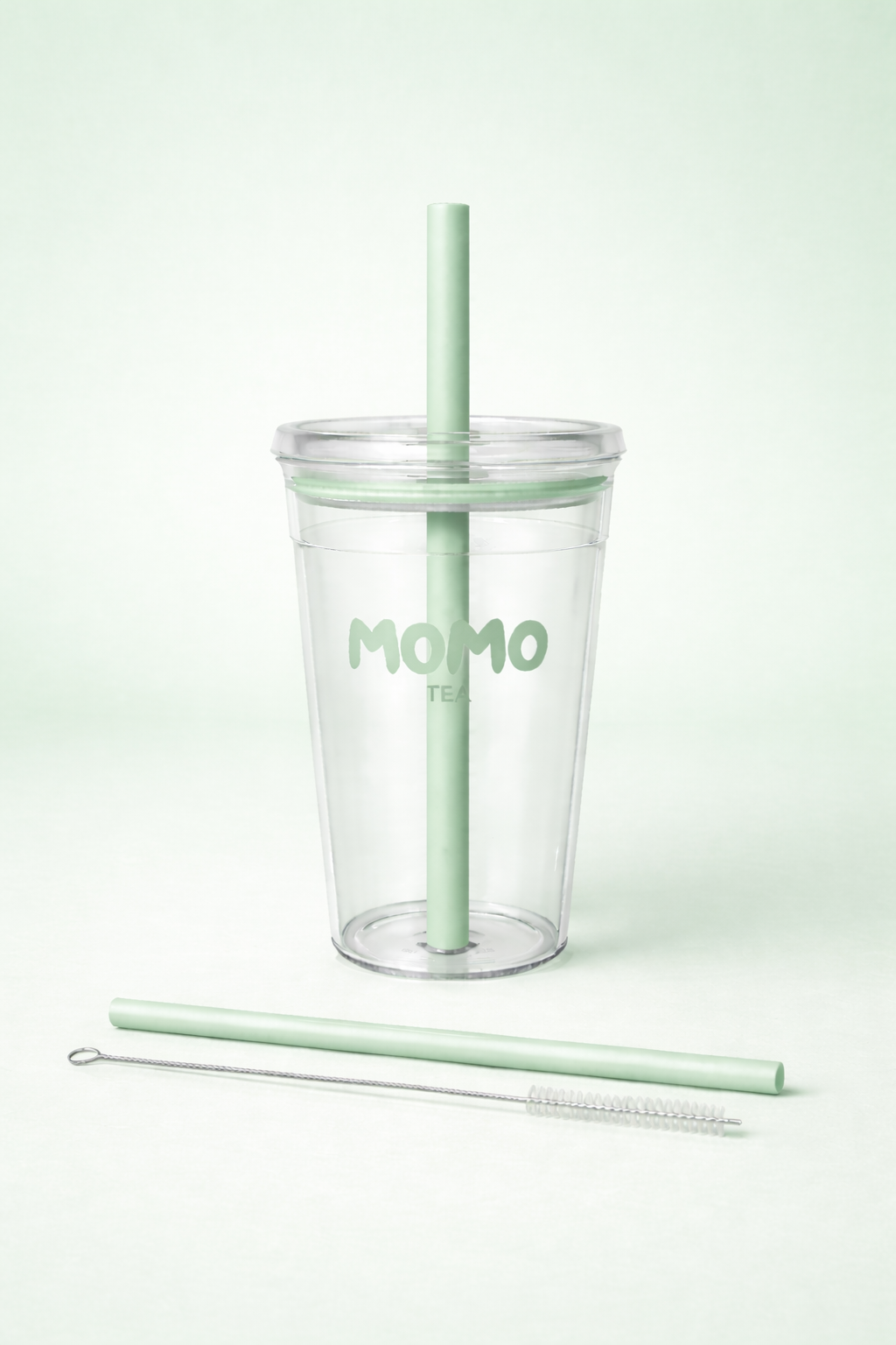 A clear plastic tumbler cup with a mint green straw and matching lid, featuring the word 'MOMO' printed on the front. A second mint green straw and a small metal cleaning brush are placed in front on a clean, light mint green background.